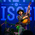 Canadian country artist Brett Kissell holding up his guitar while playing it and singing into a microphone on stage at the Algonquin Commons Theatre