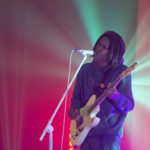 Hip hop artist Daniel Caesar playing guitar and singing with green and red lights softly lighting him from behind
