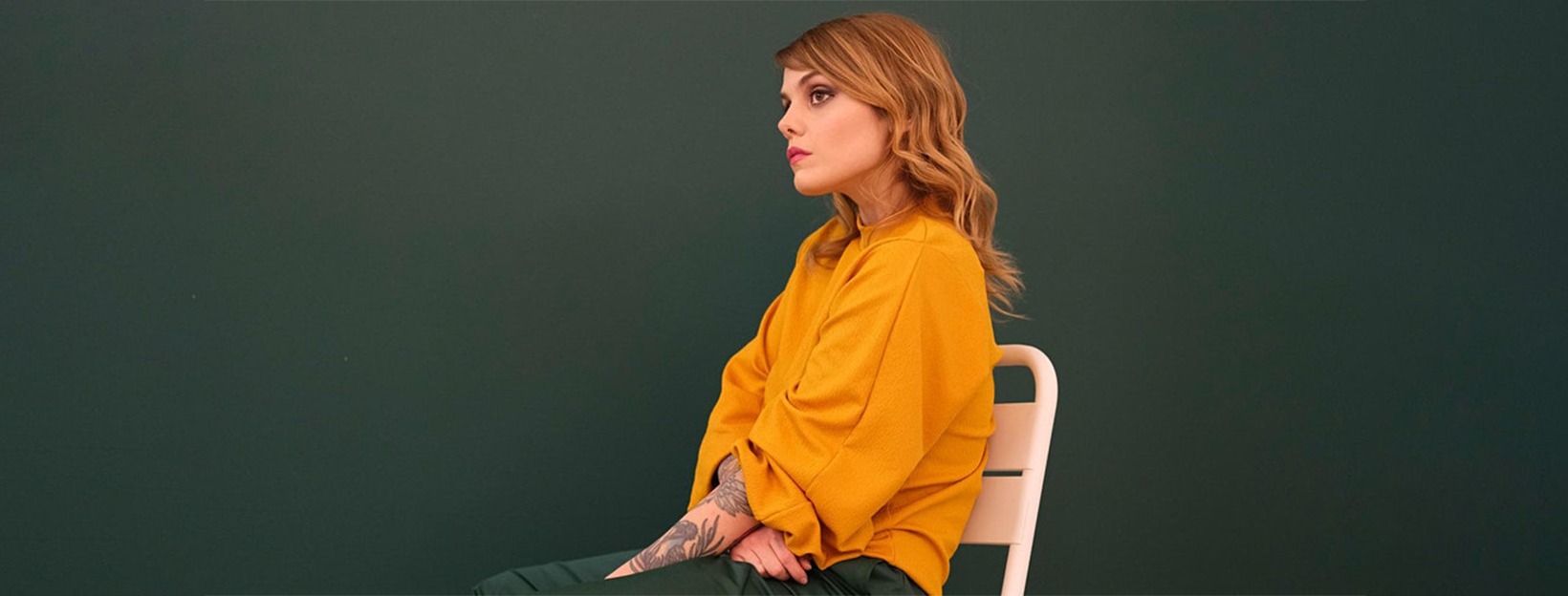 Coeur de pirate sitting in a white metal chair looking off to the distance wearing an orange sweater and green pants