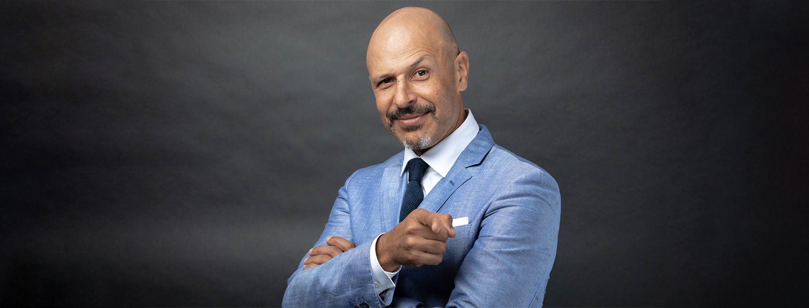 Maz Jobrani in a light blue suit pointing at the lens with a raised eyebrow